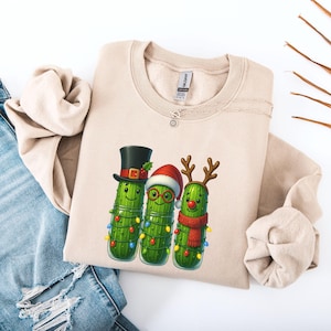 Könnte beinhalten: Ein beigefarbenes Sweatshirt mit Rundhalsausschnitt und einer Grafik von drei grünen Gewürzgurken, die für die Feiertage geschmückt sind. Eine Gurke trägt einen schwarzen Zylinder, eine eine rote Weihnachtsmütze mit Brille und eine ein Rentiergeweih.