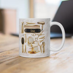 Könnte beinhalten: Ein weißer Keramikbecher mit einem goldfarbenen Grafikdruck verschiedener Friseurwerkzeuge, darunter ein Friseurstuhl, ein Haartrockner, eine Schere, ein Kamm und Bürsten. Ein schwarzes Namensschild in der Mitte zeigt den Namen Lisa in Goldschrift.