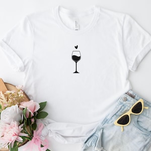 Könnte beinhalten: Ein weißes kurzärmeliges T-Shirt mit einer minimalistischen schwarzen Grafik eines Weinglases, über dessen Rand sich ein kleines Herzsymbol befindet. Das Design ist mittig auf der Brust des Kleidungsstücks platziert, das einen Rundhalsausschnitt hat.
