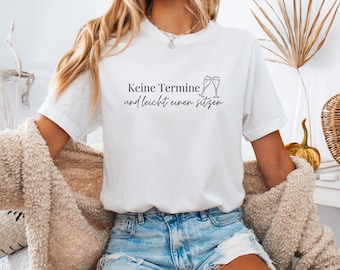 Op de afbeelding: Een wit t-shirt met korte mouwen voorzien van de zwarte tekst 'Keine Termine und leicht einen sitzen' en een afbeelding van twee klinkende champagneglazen. Het shirt heeft een ronde hals en wordt gedragen met een lichtblauwe spijkerbroek.
