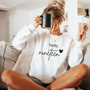 May include: A white long-sleeve crewneck sweatshirt featuring the black printed text hello nineteen with a small black heart icon. The sweatshirt has a classic fit with ribbed cuffs and a ribbed neckline, providing a clean and simple aesthetic.