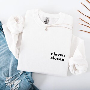 Op de afbeelding: Witte crewneck sweatshirt met de tekst "eleven eleven" in zwart. Een zilveren ketting met een kleine hanger is over de sweatshirt gedrapeerd. De sweatshirt is op een spijkerbroek met scheuren gevouwen.