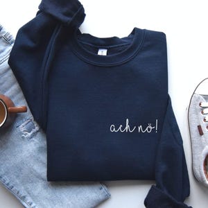 Puede incluir: Una sudadera azul marino con la frase "ach nö!" en escritura blanca. La sudadera doblada está acompañada de unos vaqueros azules, una taza marrón y una zapatilla gris. Prenda de vestir.