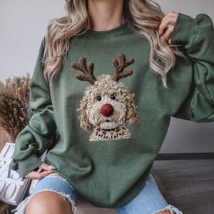 Könnte beinhalten: Ein olivgrüner Rundhals-Pullover mit einer strukturierten, gestickten Grafik eines flauschigen, hellen Hundes mit braunem Geweih und einer auffälligen roten Nase. Der Hund trägt ein kleines, gemustertes Halsband um den Hals.