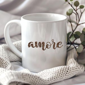 Puede incluir: Taza de cerámica blanca con la palabra "amore" en estampado de leopardo. La taza, con asa, está sobre una manta de punto blanca. Ideal para café o té.