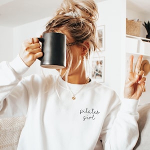 May include: A white long-sleeve crewneck sweatshirt featuring the black text "pilates girl" printed on the upper left chest area. The garment is made of a solid white fabric with a standard ribbed neckline, cuffs, and hem.