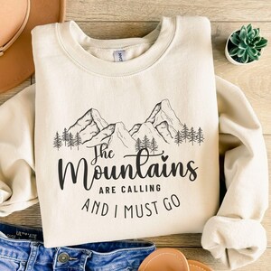 Könnte beinhalten: Cremefarbenes Sweatshirt mit schwarzer Grafik von Bergspitzen und Kiefern. Der Text lautet: "The Mountains are Calling and I Must Go". Ein lässiges, bequemes Oberteil für Outdoor-Enthusiasten.
