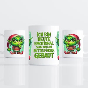 Könnte beinhalten: Ein weißer Keramikbecher mit einem grünen, mürrischen Elf-Charakter, der eine rote Weihnachtsmütze und einen Pullover trägt. In der Mitte steht der deutsche Text: ICH BIN HEUTE EMOTIONAL SEHR NAH AM MITTELFINGER GEBAUT, gedruckt in fetten grünen Buchstaben.
