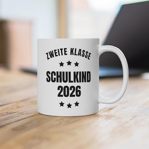 Peut inclure: Un mug en céramique blanche avec le texte noir ZWEITE KLASSE SCHULKIND 2026, disposé avec trois étoiles au-dessus et en dessous du texte central. Le mug possède une anse standard et un corps cylindrique lisse.