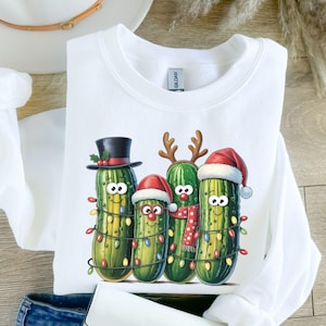 Puede incluir: Sudadera blanca con un diseño festivo de cuatro pepinillos antropomórficos. Cada pepinillo está decorado con luces navideñas y accesorios navideños, incluyendo gorros de Papá Noel, cuernos de reno y un sombrero de copa. La sudadera es de material suave.