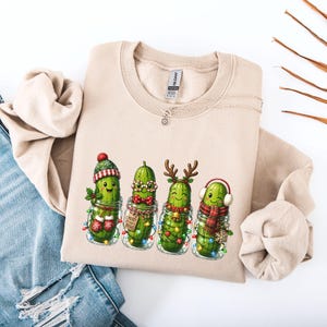 Könnte beinhalten: Ein sandfarbenes Sweatshirt mit Rundhalsausschnitt und einem Aufdruck von vier grünen Gewürzgurken in Einmachgläsern. Die Gurken sind weihnachtlich geschmückt mit einer Strickmütze, einer roten Brille, einer Fliege, Rentiergeweih und Ohrenschützern.