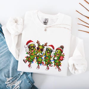 Könnte beinhalten: Ein weißes Sweatshirt mit Rundhalsausschnitt und einem Aufdruck von vier grünen Gewürzgurken in Weihnachtskleidung. Die Gurken tragen Weihnachtsmützen, Rentiergeweihe und Sonnenbrillen und sind mit bunten Lichterketten geschmückt.