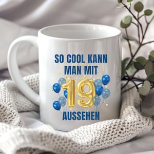May include: White ceramic mug with the text "SO COOL KANN MAN MIT 19 AUSSEHEN" in blue and gold. The number 19 is in gold, surrounded by blue balloons. The mug is on a white knitted blanket.