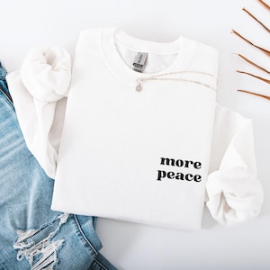 Könnte beinhalten: Ein weißes Sweatshirt mit Rundhalsausschnitt und dem schwarzen Aufdruck "more peace". Eine silberne Halskette mit kleinem Anhänger liegt über der Vorderseite. Das Sweatshirt liegt auf einer Jeans.