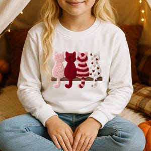 Può includere: Una felpa bianca a maniche lunghe con la grafica di quattro gatti seduti in fila su un ramo marrone. I gatti presentano texture e motivi diversi, tra cui rosa chiaro a tinta unita, rosso scuro a tinta unita, strisce rosse e rosa, e bianco con cuori rossi.