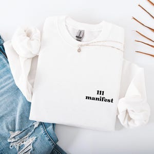 Op de afbeelding: Witte crewneck sweatshirt met de tekst "111 manifest" in zwart. Een zilveren ketting met een kleine hanger is over de sweatshirt gedrapeerd. De sweatshirt ligt op een wit oppervlak naast een spijkerbroek.