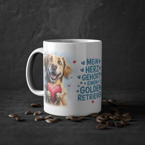 Może przedstawiać: Biały ceramiczny kubek z kolorową ilustracją golden retrievera trzymającego czerwone serce. Napis brzmi "MEIN HERZ GEHÖRT EINEM GOLDEN RETRIEVER" i jest otoczony małymi niebieskimi oraz czerwonymi ikonami serc. Kubek posiada uchwyt po lewej stronie.