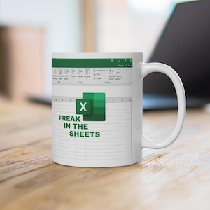 Op de afbeelding: Een witte keramische mok met een opdruk van een spreadsheet-interface. Het ontwerp bevat het groene Microsoft Excel-logo en de tekst "FREAK IN THE SHEETS" in dikgedrukte groene hoofdletters, gecentreerd over een raster van spreadsheetcellen.