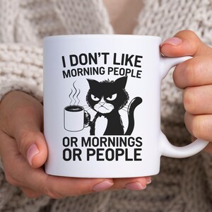 Könnte beinhalten: Ein weißer Keramikbecher mit einer schwarzen Grafik einer mürrischen Katze, die eine dampfende Tasse Kaffee hält. Der in Schwarz gedruckte Text lautet: I DON'T LIKE MORNING PEOPLE OR MORNINGS OR PEOPLE. Der Becher hat einen geschwungenen Henkel.
