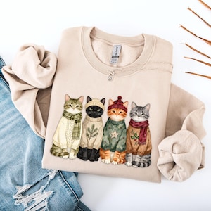 Pode incluir: Uma camisola bege de gola redonda com uma impressão gráfica de quatro gatos a usar camisolas, cachecóis e gorros de inverno. Os gatos estão representados em várias cores com padrões festivos como ramos de pinheiro e estrelas nas malhas.