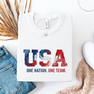 Könnte beinhalten: Ein weißes T-Shirt mit Rundhalsausschnitt und dem Schriftzug USA in einer rot-weiß-blauen Pinselstrich-Schriftart. Unter den Buchstaben steht der Text ONE NATION. ONE TEAM. in dunkelblauen und roten serifenlosen Großbuchstaben.