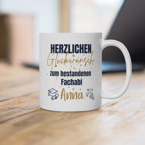 Könnte beinhalten: Eine weiße Keramiktasse mit dem Text "HERZLICHEN Glückwunsch zum bestandenen Fachabi Anna" in dunkelblauer und goldener Schrift. Das Design enthält goldene Konfetti-Akzente, eine kleine Doktorhut-Grafik, ein Diplom und ein Party-Popper-Symbol.