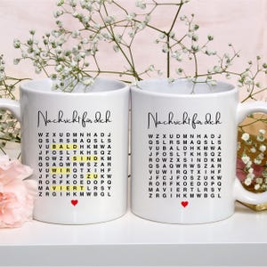 May include: Two white ceramic mugs with black handles. Each mug has a black and white word search puzzle with the words "Nachricht für dich" (German for "message for you") at the top. The word "SIND" (German for "are") is highlighted in yellow on each mug. A small red heart is at the bottom of each mug.