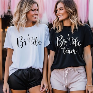Op de afbeelding: Twee vrouwen dragen bijpassende t-shirts. Eén shirt is wit met het woord "Bride" en een handgebaar. Het andere shirt is zwart met de woorden "Bride Team" en een handgebaar. De shirts hebben korte mouwen.