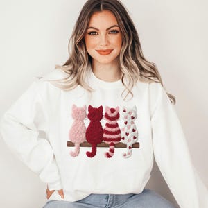 Op de afbeelding: Een wit sweatshirt met ronde hals en een afbeelding van vier pluizige katten die op een bruine tak zitten. De katten zijn lichtroze, effen donkerrood, rood-wit gestreept en wit met kleine rode hartjes, allemaal van de achterkant gezien.