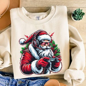 Op de afbeelding: Crèmekleurige sweatshirt met een afbeelding van de Kerstman met een koptelefoon en zonnebril, die een gamecontroller vasthoudt. De Kerstman draagt een rood pak met witte randen, en het ontwerp bevat hulst en bessen.