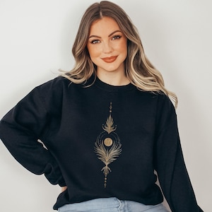 May include: A black long-sleeve crewneck sweatshirt featuring a centered gold-colored graphic design. The print consists of a vertical arrangement of fine lines, delicate leaf-like patterns, and a central circular sun or moon motif.