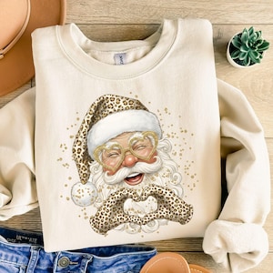 Può includere: Una felpa girocollo color crema con una stampa di Babbo Natale che indossa un cappello leopardato e occhiali dorati a forma di cuore. Babbo Natale sorride e forma un cuore con le mani guantate di leopardato, circondato da glitter dorati.