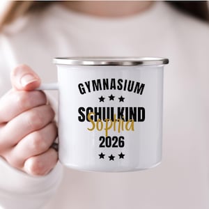 Könnte beinhalten: Weißer Emaillebecher mit silbernem Rand und Henkel. Der Becher trägt die Worte "GYMNASIUM SCHULKIND Sophia 2026" in schwarzer und goldener Schrift mit Sternen. Der Becher wird von einer Person gehalten.