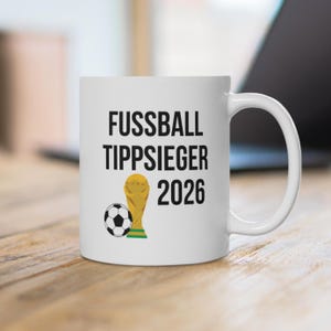 May include: A white ceramic mug featuring the black text FUSSBALL TIPPSIEGER 2026, a black and white football, and a gold-coloured trophy illustration. The mug has a standard handle and a smooth, cylindrical body design for holding beverages.