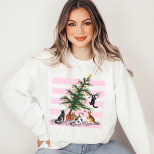 Könnte beinhalten: Ein weißes Sweatshirt mit Rundhalsausschnitt und einem Grafikdruck eines grünen Weihnachtsbaums, der mit bunten Kugeln und einem gelben Stern geschmückt ist. Vier Katzen sind um den Baum herum platziert, eine davon klettert, vor einem rosa gestreiften Hintergrund.