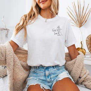 Könnte beinhalten: Ein weißes Kurzarm-T-Shirt mit einer schwarzen Strichzeichnung auf der linken Brust. Die Grafik zeigt eine Hand, die ein Weinglas hält, mit einer Herzform darüber und dem Text "Wine Time" in kursiver Schrift unter dem Glas.