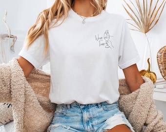 May include: A white short-sleeve t-shirt featuring a black line art graphic on the left chest. The graphic depicts a hand holding a wine glass with a heart shape above it and the text "Wine Time" written in a cursive script font below the glass.