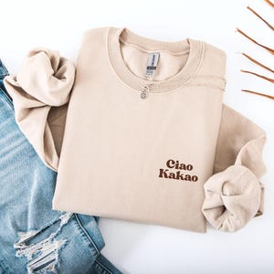 Könnte beinhalten: Beiges Sweatshirt mit dem Text "Ciao Kakao" in braunen Buchstaben. Eine silberne Halskette mit einem kleinen Anhänger liegt auf dem Sweatshirt. Das Sweatshirt ist auf einer Jeans gefaltet.