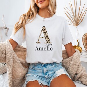 Könnte beinhalten: Ein weißes kurzärmeliges T-Shirt mit einem großen Buchstaben A in braun-schwarzem Leopardenmuster. Unter dem Buchstaben steht der Text est. 2011 und der Name Amelie in einer schwarzen Serifenschrift. Das T-Shirt wird mit hellblauen Jeansshorts getragen.