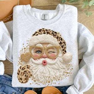 Op de afbeelding: Een lichtgrijs sweatshirt met ronde hals en een centrale afbeelding van een knipogende kerstman met een muts met luipaardprint en een goudkleurige bril. Het ontwerp bevat een gestructureerde witte baard en gouden glitterdetails.