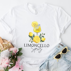 Op de afbeelding: Wit T-shirt met een aquarel illustratie van een limoncello spritz cocktail, citroenen en groen. De woorden "Limoncello Spritz" staan eronder in zwart schrift.
