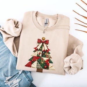 Könnte beinhalten: Ein beigefarbenes Sweatshirt mit Rundhalsausschnitt und einer Grafik eines gesteppten Weihnachtsbaums. Der Baum besteht aus roten, grünen und cremefarbenen Stoffstücken mit goldenen Knöpfen, einer roten Schleife und einem goldenen Stern. Eine Silberkette ist sichtbar.
