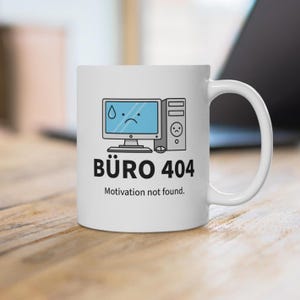 Könnte beinhalten: Ein weißer Keramikbecher mit einer Grafik eines blauen Computermonitors mit traurigem Gesicht und einem grauen Computergehäuse. Unter der Grafik steht der Text BÜRO 404 und Motivation not found. Der Becher hat einen Standardhenkel und einen zylindrischen Körper.