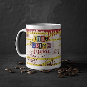Könnte beinhalten: Ein weißer Keramikbecher mit einem grafischen Design, das rote Bluttropfen am oberen Rand, gelbes Polizeiabsperrband und eine Collage aus Zeitungsausschnitten zeigt. Der Text True Crime Junkie ist in einer Lösegeldbrief-Schriftart gedruckt.