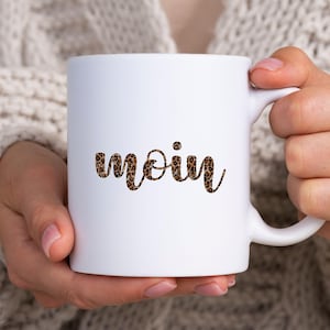 May include: White ceramic mug with the word "moin" in a leopard print design. The mug has a curved handle and is held by a person. The mug is a standard size for coffee or tea.