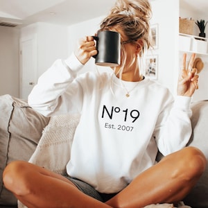 May include: A white long-sleeve crewneck sweatshirt featuring the black text N°19 and Est. 2007 printed on the center of the chest. The garment has a classic fit with ribbed cuffs and a ribbed neckline, providing a clean and simple aesthetic.
