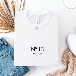 Könnte beinhalten: Ein weißes T-Shirt mit Rundhalsausschnitt und dem schwarzen Aufdruck "N°13" sowie "Est. 2013" auf der Vorderseite. Das Shirt hat einen Standardkragen und ein sichtbares Markenetikett im Nackenbereich. Das Design ist mittig platziert.
