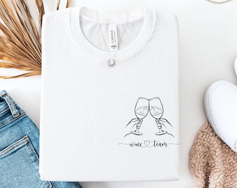 May include: A white t-shirt featuring a black line art graphic of two hands clinking wine glasses together. Below the glasses, the text reads wine team with a small heart symbol connecting the two words in a cursive script font.