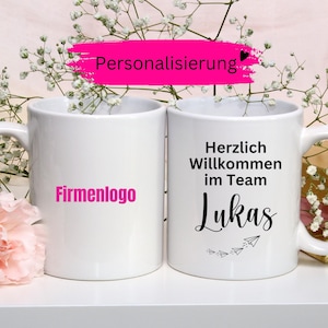 May include: Two white ceramic mugs. One mug has the text "Firmenlogo" in pink. The other mug has the text "Herzlich Willkommen im Team Lukas" in black, with paper airplane graphics. The word "Personalisierung" is in pink.