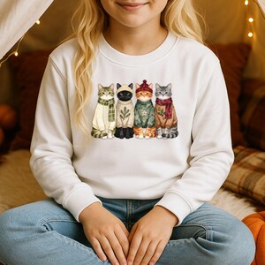 Op de afbeelding: Een wit sweatshirt met lange mouwen en een grafische print van vier katten die op een rij zitten. Elke kat draagt een gebreide trui in een andere kleur en een sjaal of muts, in groen-, crème- en bruintinten met feestelijke patronen.